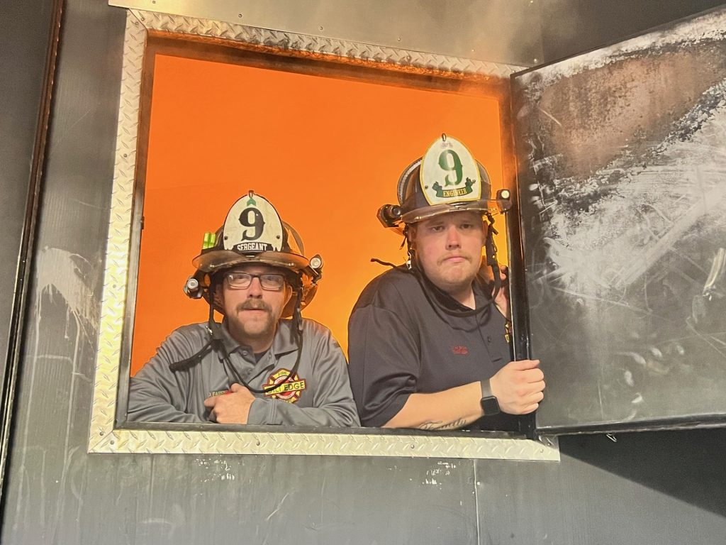 Two firefighters wearing helmets labeled "9" look out of a square, metal-framed window with an orange background behind them and a partially open, scorched metal door to the right.